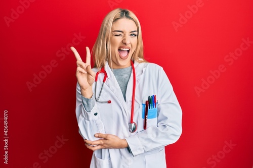 Young caucasian woman wearing doctor uniform and stethoscope smiling with happy face winking at the camera doing victory sign. number two.