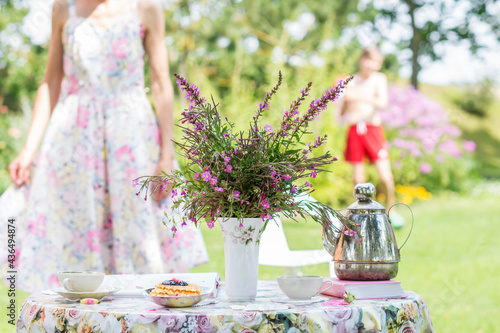 Wild flowers bouquet of  rosebay willowherbs or  fireweed in a vase on a flowery tablecloth , cake and teapot , cups with tea, woman and boy standing in background outdoors in summer time in garden