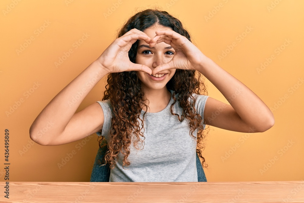 Teenager hispanic girl wearing casual clothes sitting on the table doing heart shape with hand and fingers smiling looking through sign Teenager hispanic girl wearing casual clothes sitting on the table doing heart shape with hand and fingers smiling looking through sign