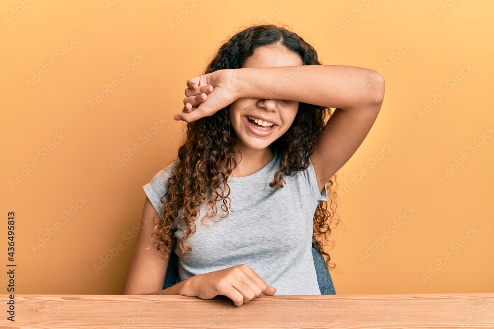 Teenager hispanic girl wearing casual clothes sitting on the table covering eyes with arm smiling cheerful and funny. blind concept. Teenager hispanic girl wearing casual clothes sitting on the table covering eyes with arm smiling cheerful and funny. blind concept.