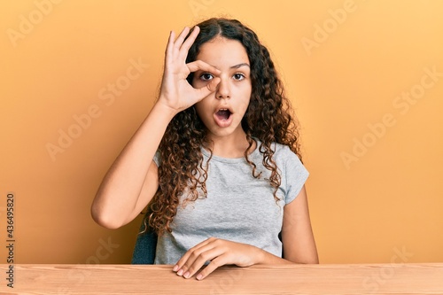 Teenager hispanic girl wearing casual clothes sitting on the table doing ok gesture shocked with surprised face, eye looking through fingers. unbelieving expression. © Krakenimages.com