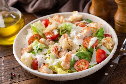 Salad with grilled chicken breast, romaine lettuce, tomatoes and sauce known as Caesar salad served on plate, wooden table background