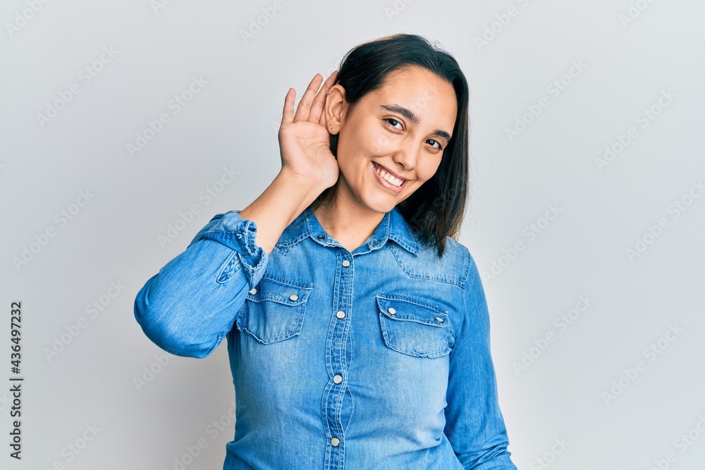 Young hispanic woman wearing casual denim jacket smiling with hand over ear listening an hearing to rumor or gossip. deafness concept. Young hispanic woman wearing casual denim jacket smiling with hand over ear listening an hearing to rumor or gossip. deafness concept.