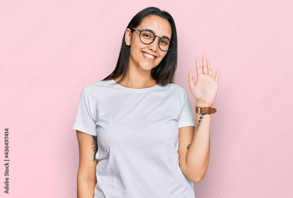 Young hispanic woman wearing casual white t shirt waiving saying hello happy and smiling, friendly welcome gesture Young hispanic woman wearing casual white t shirt waiving saying hello happy and smiling, friendly welcome gesture