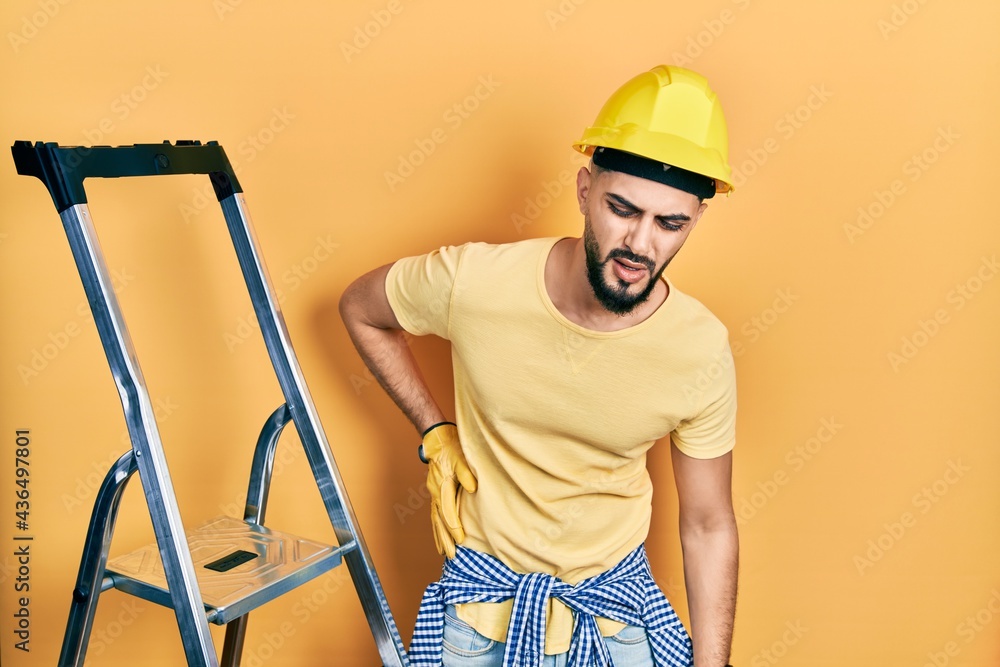 Handsome man with beard by construction stairs wearing hardhat suffering of backache, touching back with hand, muscular pain Handsome man with beard by construction stairs wearing hardhat suffering of backache, touching back with hand, muscular pain