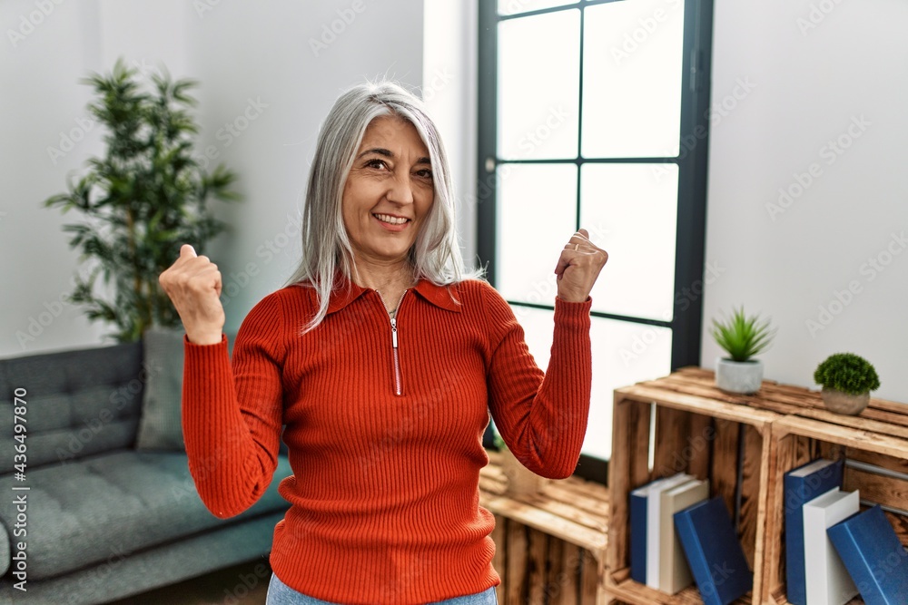 Middle age grey-haired woman wearing casual clothes standing at home very happy and excited doing winner gesture with arms raised, smiling and screaming for success. celebration concept. Middle age grey-haired woman wearing casual clothes standing at home very happy and excited doing winner gesture with arms raised, smiling and screaming for success. celebration concept.