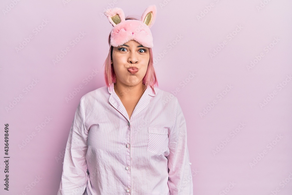 Hispanic woman with pink hair wearing sleep mask and pajama puffing cheeks with funny face. mouth inflated with air, crazy expression. Hispanic woman with pink hair wearing sleep mask and pajama puffing cheeks with funny face. mouth inflated with air, crazy expression.