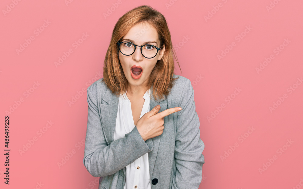 Young caucasian woman wearing business style and glasses surprised pointing with finger to the side, open mouth amazed expression. Young caucasian woman wearing business style and glasses surprised pointing with finger to the side, open mouth amazed expression.