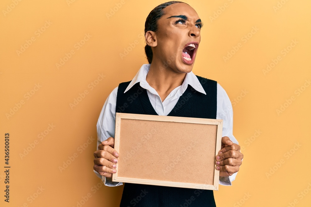 Hispanic man wearing make up and long hair holding empty corkboard angry and mad screaming frustrated and furious, shouting with anger. rage and aggressive concept. Hispanic man wearing make up and long hair holding empty corkboard angry and mad screaming frustrated and furious, shouting with anger. rage and aggressive concept.