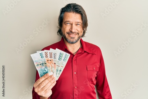 Middle age handsome man holding czech koruna banknotes looking positive and happy standing and smiling with a confident smile showing teeth