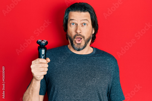 Middle age caucasian man holding electric razor machine scared and amazed with open mouth for surprise, disbelief face