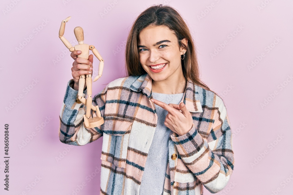 Young caucasian girl holding small wooden manikin smiling happy pointing with hand and finger Young caucasian girl holding small wooden manikin smiling happy pointing with hand and finger
