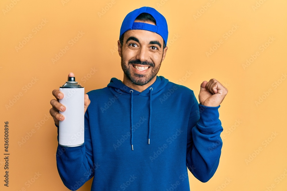 Young hispanic man wearing sweatshirt holding graffiti spray screaming proud, celebrating victory and success very excited with raised arm