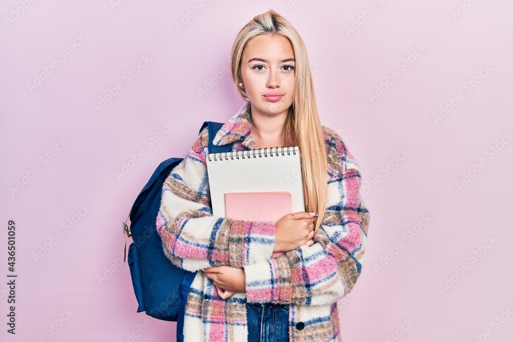 Young blonde girl holding student backpack and books relaxed with serious expression on face. simple and natural looking at the camera. Young blonde girl holding student backpack and books relaxed with serious expression on face. simple and natural looking at the camera.