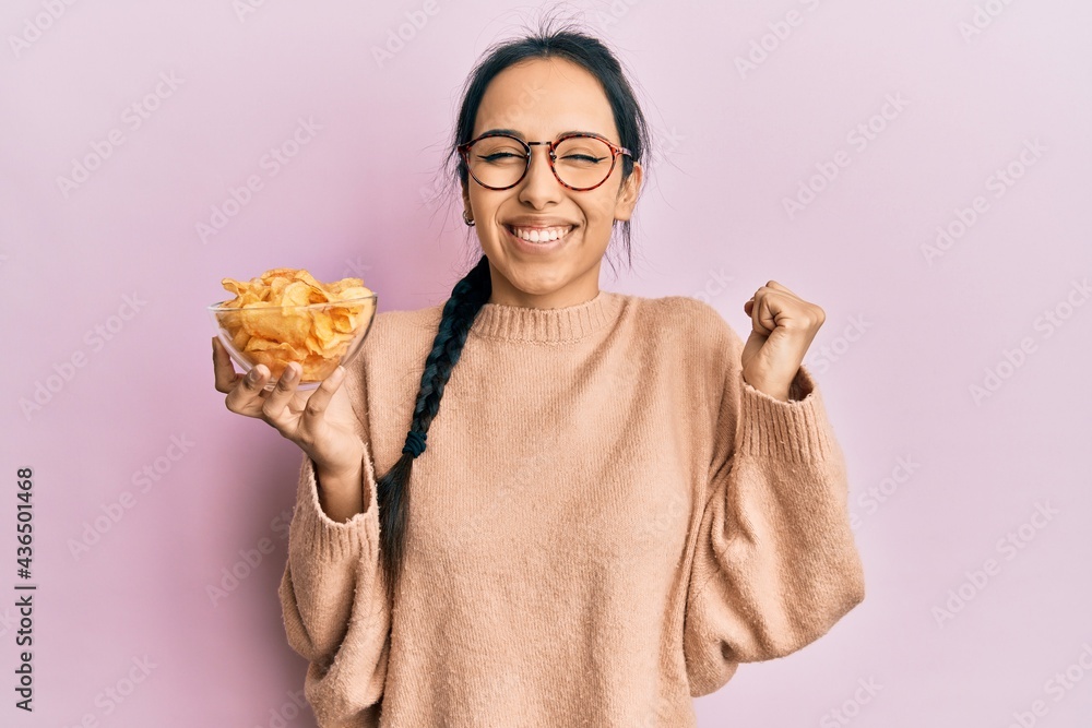 Young hispanic girl holding potato chip screaming proud, celebrating victory and success very excited with raised arm Young hispanic girl holding potato chip screaming proud, celebrating victory and success very excited with raised arm