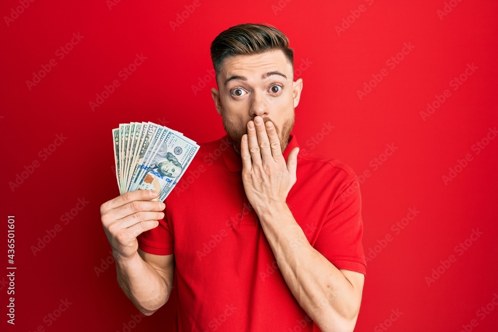 Young redhead man holding dollars covering mouth with hand, shocked and afraid for mistake. surprised expression Young redhead man holding dollars covering mouth with hand, shocked and afraid for mistake. surprised expression