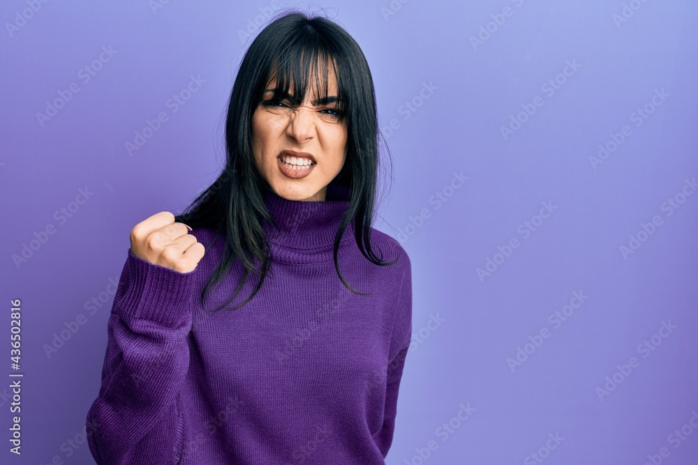 Young brunette woman with bangs wearing turtleneck sweater angry and mad raising fist frustrated and furious while shouting with anger. rage and aggressive concept. Young brunette woman with bangs wearing turtleneck sweater angry and mad raising fist frustrated and furious while shouting with anger. rage and aggressive concept.