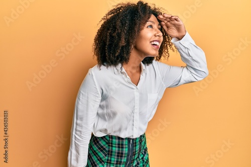 Beautiful african american woman with afro hair wearing scholar skirt very happy and smiling looking far away with hand over head. searching concept. © Krakenimages.com