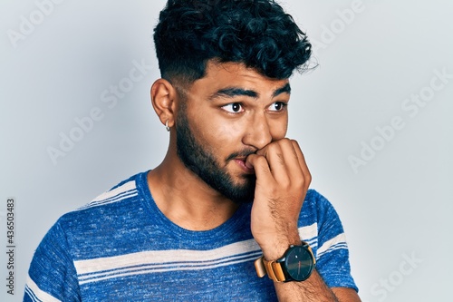 Arab man with beard wearing casual striped t shirt looking stressed and nervous with hands on mouth biting nails. anxiety problem.