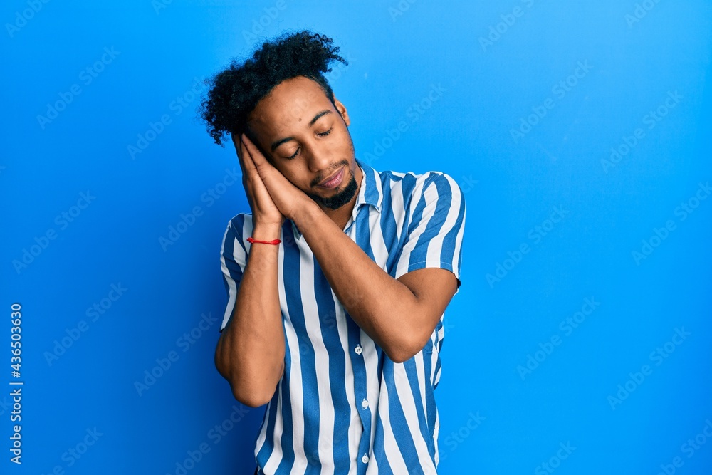 Young african american man with beard wearing casual striped shirt sleeping tired dreaming and posing with hands together while smiling with closed eyes.