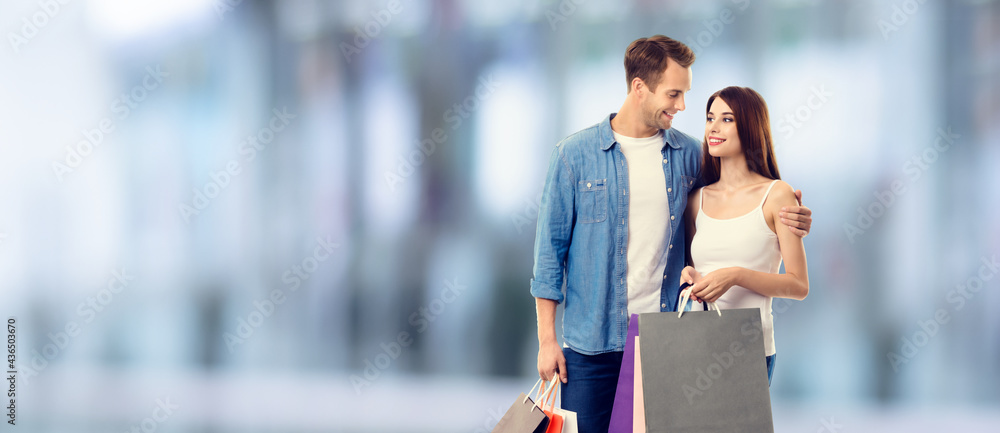 Love, holiday sales, shop, retail, consumer concept - happy couple with shopping bags, looking at each other. Standing against blurred modern interior or mall background.