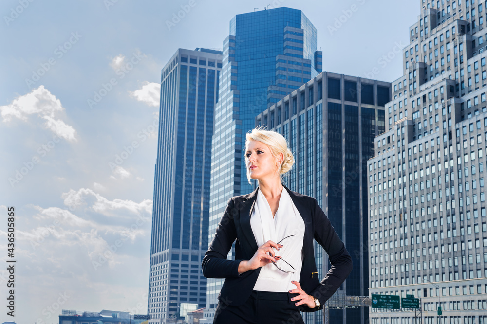Dressing in white underclothes and a black suit unbuttoned, holding glasses, a young pretty blonde businesswoman is standing in the front of a business district and hopefully looking forward.. Dressing in white underclothes and a black suit unbuttoned, holding glasses, a young pretty blonde businesswoman is standing in the front of a business district and hopefully looking forward..