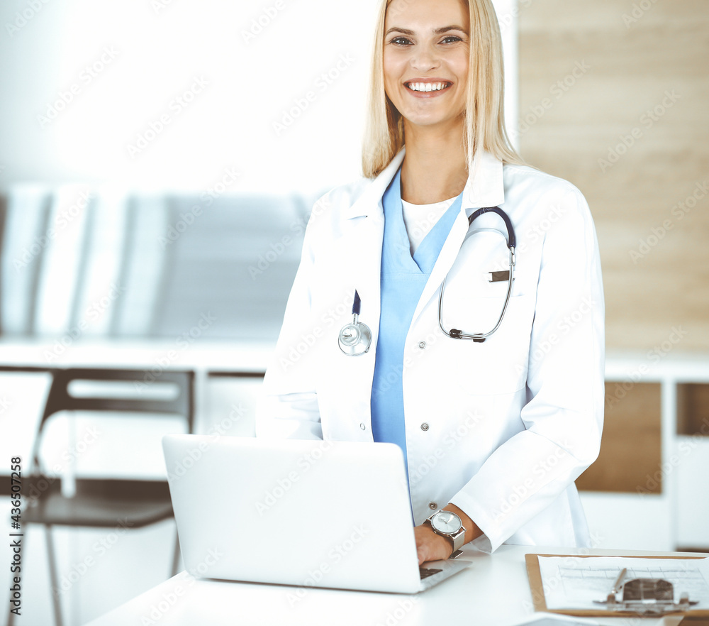 Blond female physician is lsmiling while using laptop computer. Woman-doctor at work in clinic excited and happy of her profession. Medicine concept Blond female physician is lsmiling while using laptop computer. Woman-doctor at work in clinic excited and happy of her profession. Medicine concept