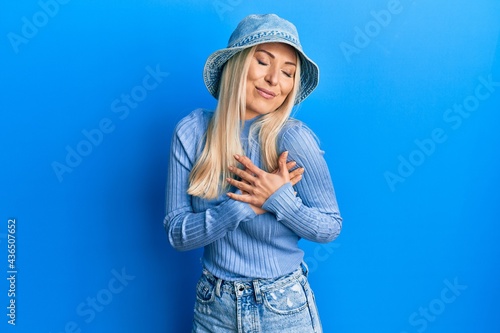 Young blonde woman wearing casual denim hat smiling with hands on chest with closed eyes and grateful gesture on face. health concept.