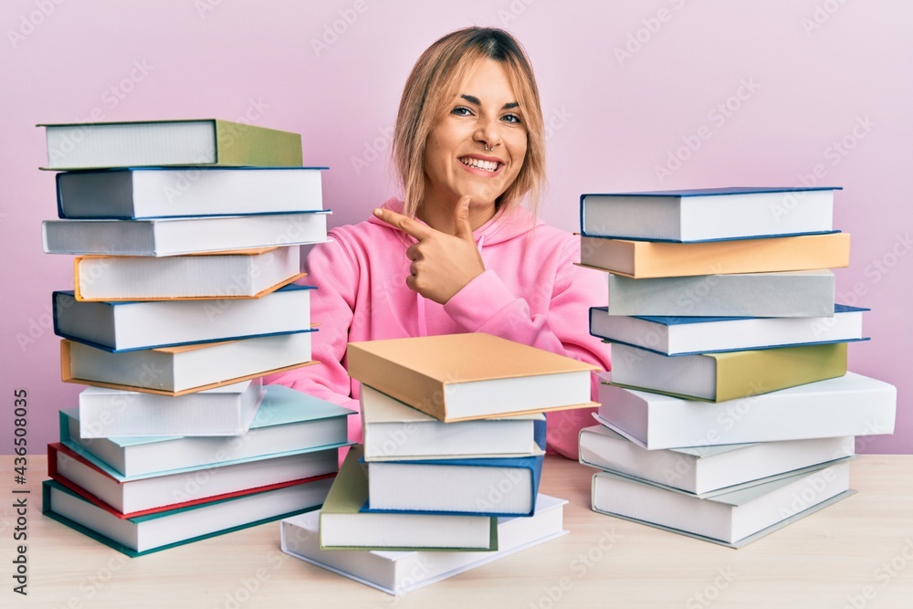 Young caucasian woman sitting on the table with books cheerful with a smile on face pointing with hand and finger up to the side with happy and natural expression Young caucasian woman sitting on the table with books cheerful with a smile on face pointing with hand and finger up to the side with happy and natural expression