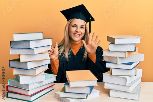 Young caucasian woman wearing graduation ceremony robe sitting on the table showing and pointing up with fingers number six while smiling confident and happy.