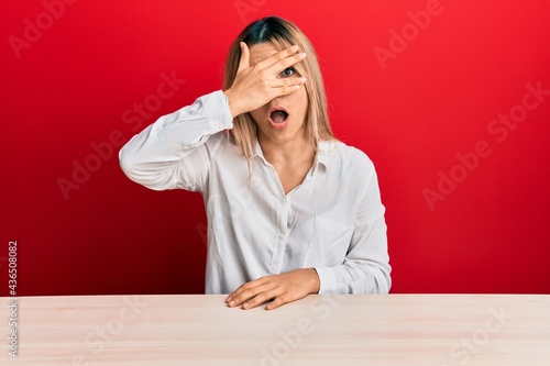 Young caucasian woman wearing casual clothes sitting on the table peeking in shock covering face and eyes with hand, looking through fingers with embarrassed expression.