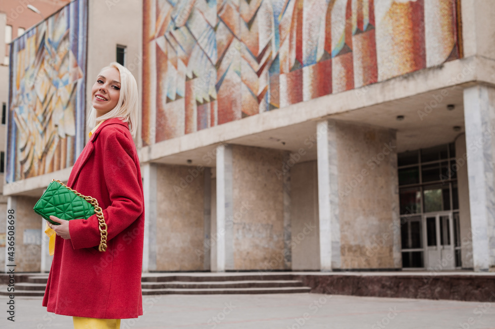 Outdoor portrait of happy smiling fashionable woman wearing bright colorful outfit with red coat, holding green leather quilted bag, posing in street of city. Copy, empty space for text