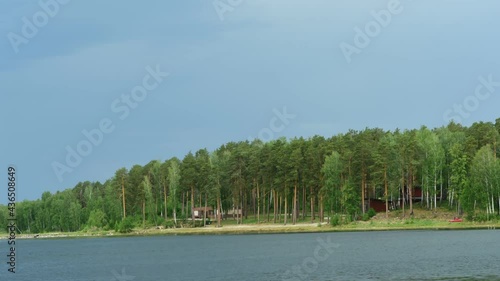 Green forest with cottage houses on the shore under a dark stormy sky before the rain. Lake Uvildy, Russia. Dramatic cloudscape. Travel, sailing, summer vacation concepts photo