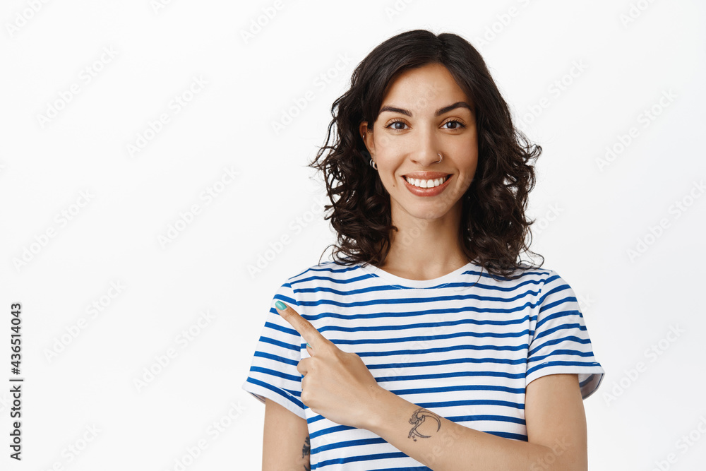 Smiling modern brunette woman pointing finger at upper left corner, showing advertisement banner on copy space, standing in striped t-shirt against white background Smiling modern brunette woman pointing finger at upper left corner, showing advertisement banner on copy space, standing in striped t-shirt against white background