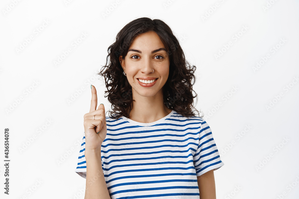 Portrait of smiling brunette woman pointing finger up, showing advertisement or logo, recommend click on link, standing in t-shirt against white background Portrait of smiling brunette woman pointing finger up, showing advertisement or logo, recommend click on link, standing in t-shirt against white background