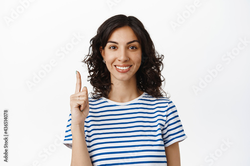 Portrait of smiling brunette woman pointing finger up, showing advertisement or logo, recommend click on link, standing in t-shirt against white background