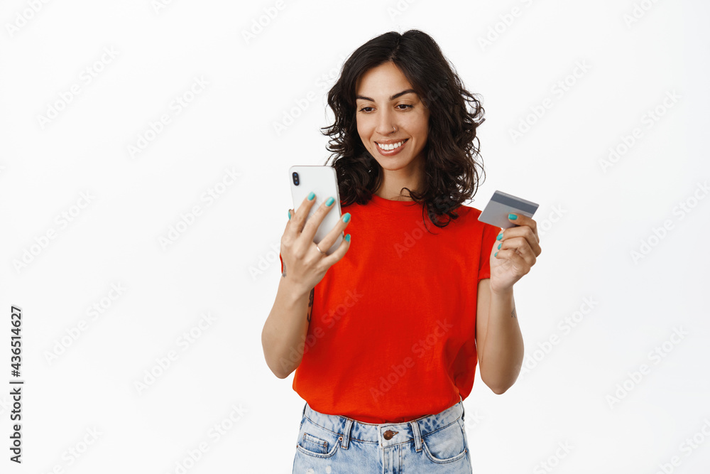 Young woman paying with smartphone, looking at phone to unlock process payment, holding credit card and mobile, standing over white background Young woman paying with smartphone, looking at phone to unlock process payment, holding credit card and mobile, standing over white background