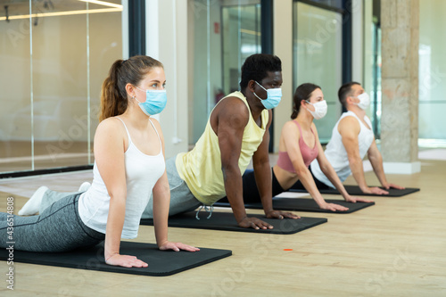 Group of sporty people in protective masks exercising pilates class in modern fitness center. Focus on young brunette. Healthy lifestyle and pandemic precautions