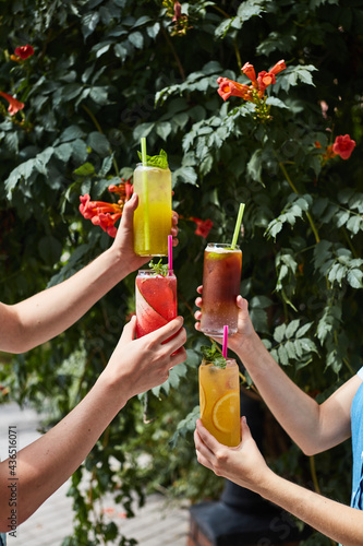 hands of guys and girls with refreshing cocktails from fruits on the background of a green hedge with red flowers 