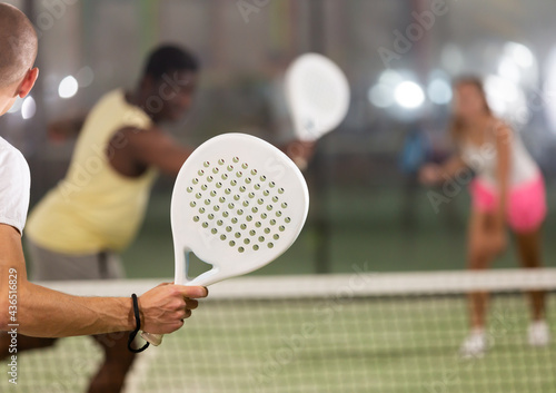 Rear view of man with white racket playing padel tennis with friends at court