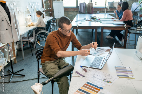 Young disable man sitting by table and working over fashion collection in large workshop photo