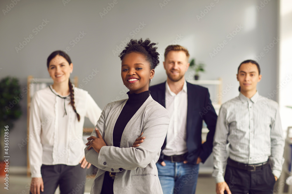 Happy aspiring black business woman in suit smiling at camera standing arms folded with team of workers behind. Portrait of ambitious female leader, executive, bank staff manager. Blurred background Happy aspiring black business woman in suit smiling at camera standing arms folded with team of workers behind. Portrait of ambitious female leader, executive, bank staff manager. Blurred background