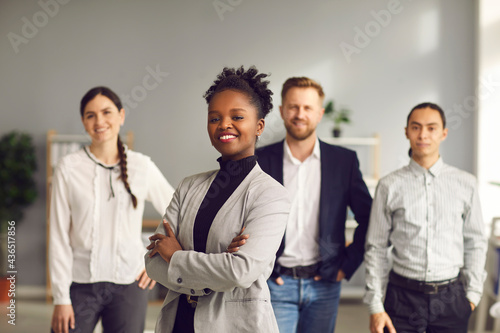 Happy aspiring black business woman in suit smiling at camera standing arms folded with team of workers behind. Portrait of ambitious female leader  executive  bank staff manager. Blurred background
