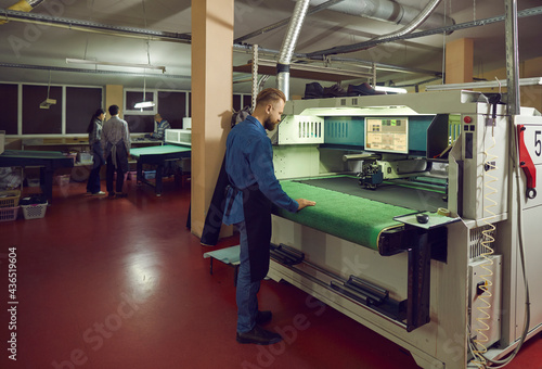 Male worker using specialized equipment standing in workshop at shoe making factory. Man working with fabric and leather cutting machine. Footwear manufacturing industry and production process concept