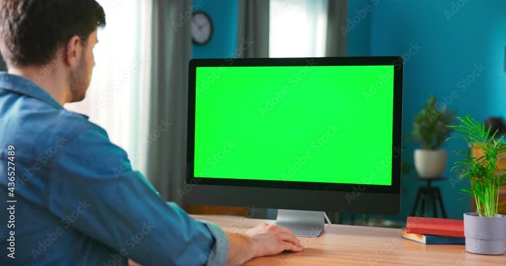 Over the Shoulder Shot of Mans Hands Typing on a keyboard computer with Green Mock-up Screen. In the Background Cozy Living Room.