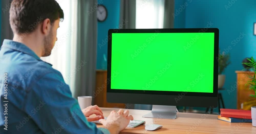 Over the Shoulder Shot of Man Typing on a keyboard computer with blank screen. Zoom in On a Modern Personal Computer with Mock-up Green Screen Display Standing on the Desk of the Cozy Home Over the Shoulder Shot of Man Typing on a keyboard computer with blank screen. Zoom in On a Modern Personal Computer with Mock-up Green Screen Display Standing on the Desk of the Cozy Home