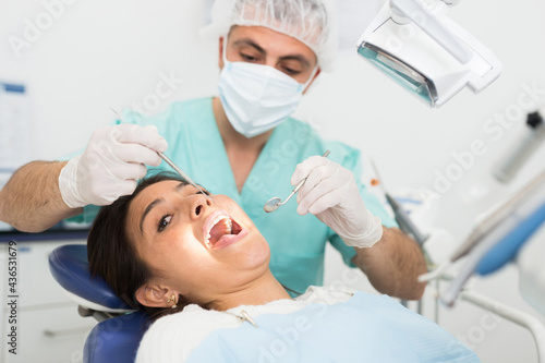 Dentist man examining a latin female patient teeth with dental tools - mirror and probe