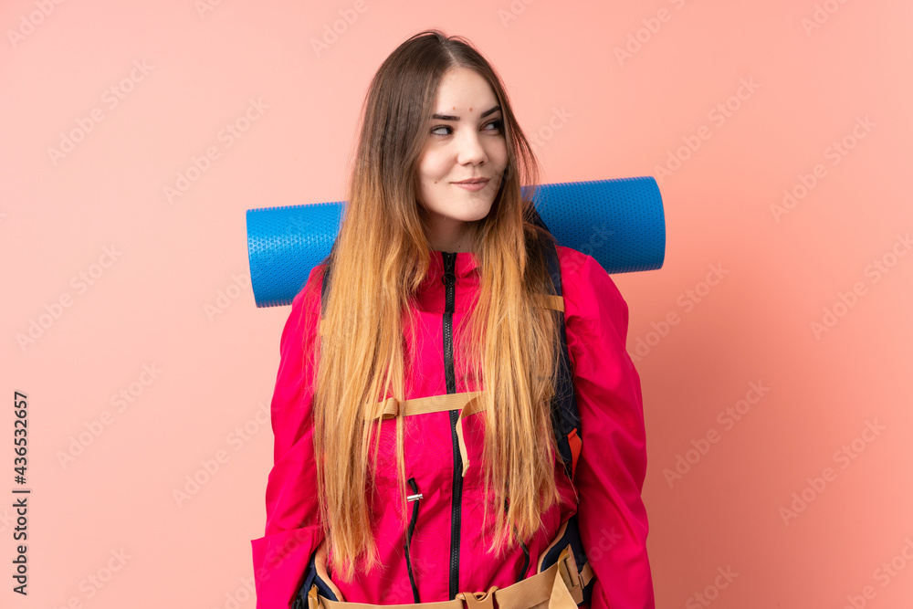 Young mountaineer girl with a big backpack isolated on pink background standing and looking to the side Young mountaineer girl with a big backpack isolated on pink background standing and looking to the side