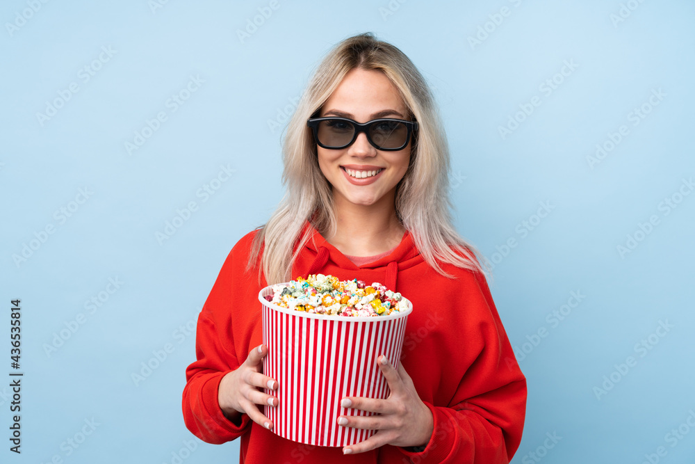Teenager girl over isolated blue background with 3d glasses and holding a big bucket of popcorns
