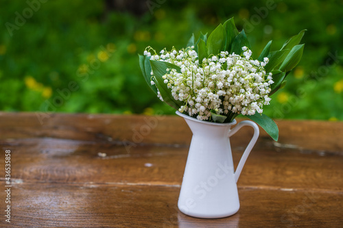 Beautiful bouquet of lilies of the valley in a white enamel vase on the wooden table in the garden